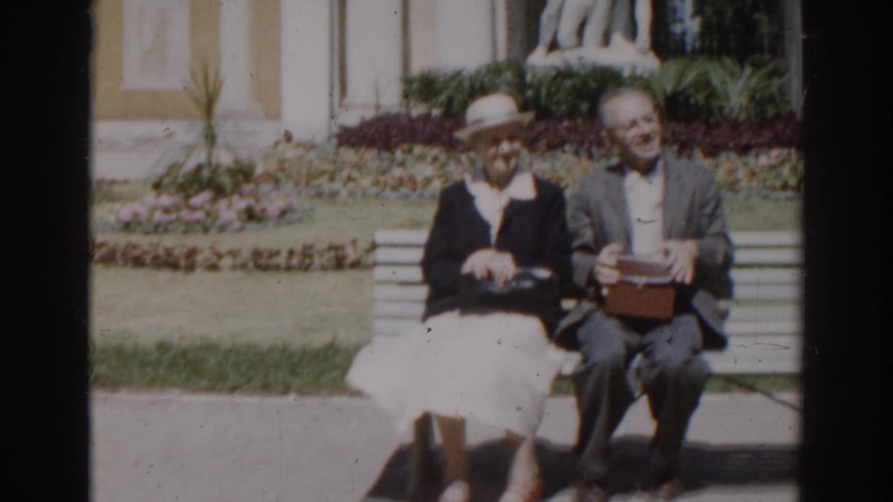 In supplied 1968 Moscow footage, an elderly couple sits together on a garden bench near flowerbeds and a pale monument facade.