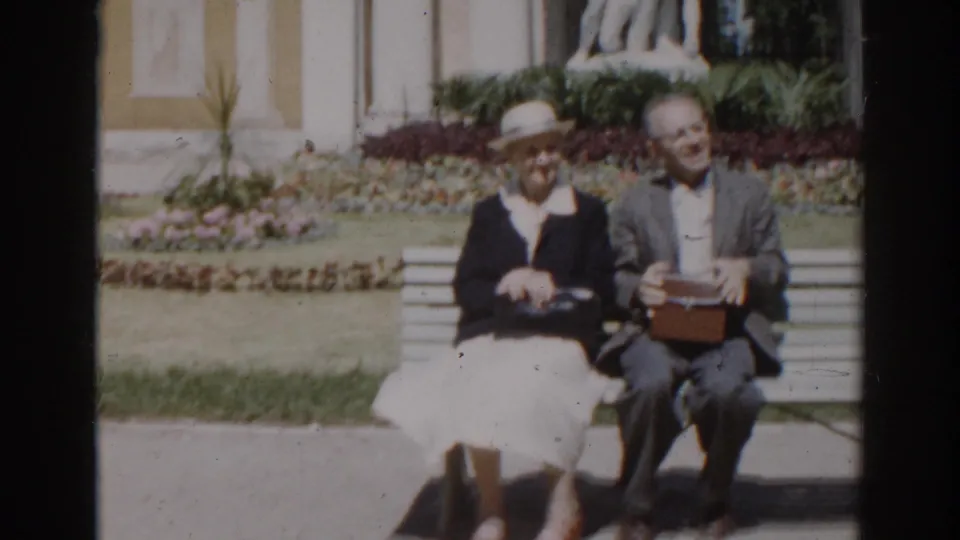 In supplied 1968 Moscow footage, an elderly couple sits together on a garden bench near flowerbeds and a pale monument facade.