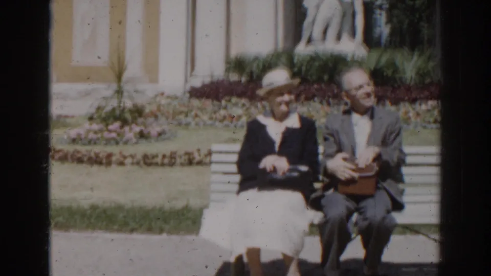 The elderly couple remains seated in the Moscow garden scene, the man holding a small box while flowerbeds and facade details sit behind them.