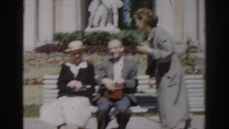 A woman in a long coat approaches the seated elderly couple in the supplied 1968 Moscow garden footage.