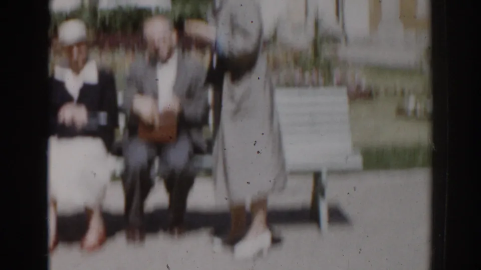 The woman stands near the seated couple in the Moscow park scene, with the bench, lawn, and flowerbeds still visible around them.