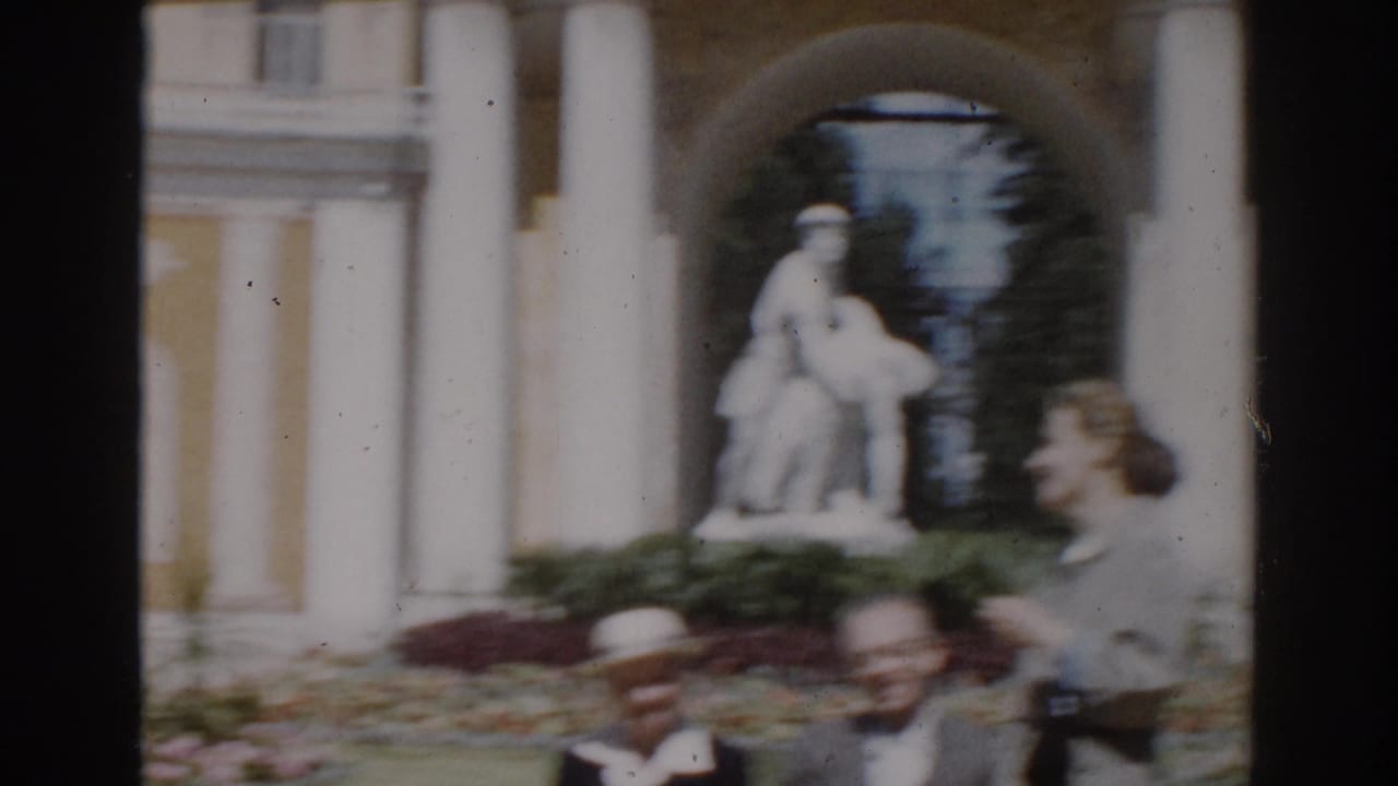 1968 Moscow home-movie frame showing visitors near a yellow columned building with a statue visible through an arched entry.