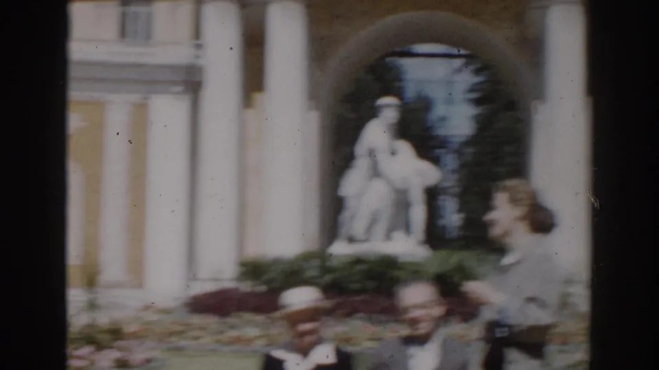 1968 Moscow home-movie frame showing visitors near a yellow columned building with a statue visible through an arched entry.