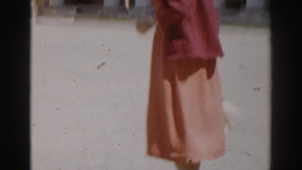 1968 Moscow home-movie frame of a woman in an orange dress standing in a sunlit courtyard beside the yellow building.