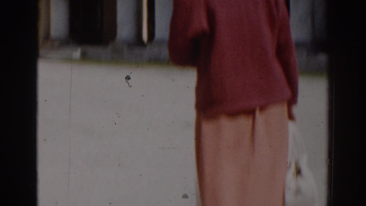 In 1968 Moscow home-movie footage, a woman in a red sweater and peach skirt walks through a sunlit courtyard.