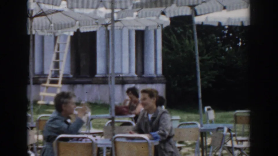 Outdoor café patrons sit at tables beneath striped umbrellas, with a columned building in the background in Moscow footage.