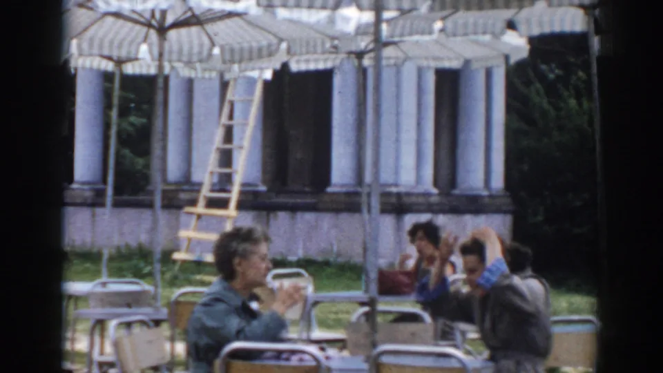 A handheld 8mm view shows café patrons at outdoor tables, striped umbrellas, grass, and a columned building in Moscow.