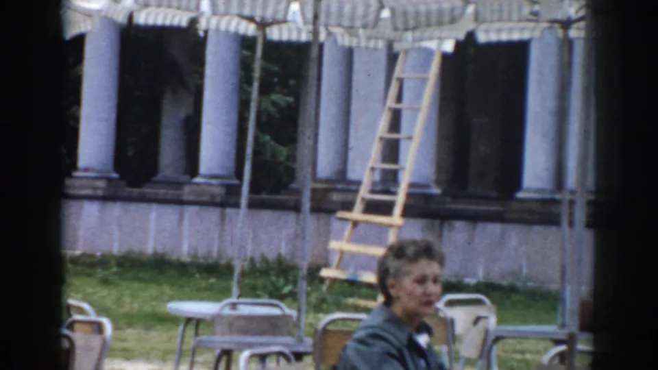 A woman appears near empty café chairs and striped umbrellas, with a columned building and ladder visible behind her.