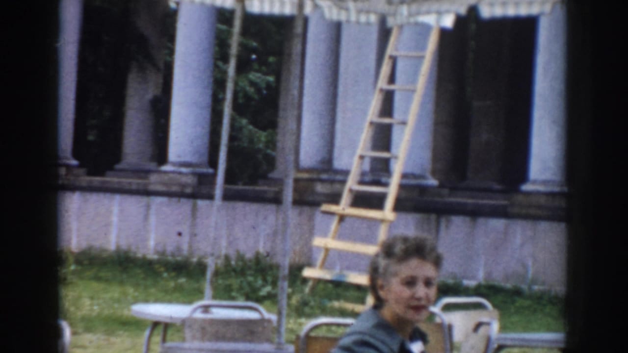1968 Moscow home-movie frame showing a woman in a green jacket by outdoor seating, with a ladder leaning near classical columns.