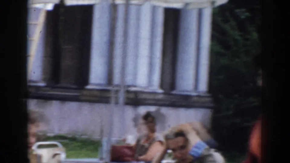 Adults sit at outdoor tables in Moscow, Russia, with a columned building, awnings, and greenery visible in soft 8mm footage.