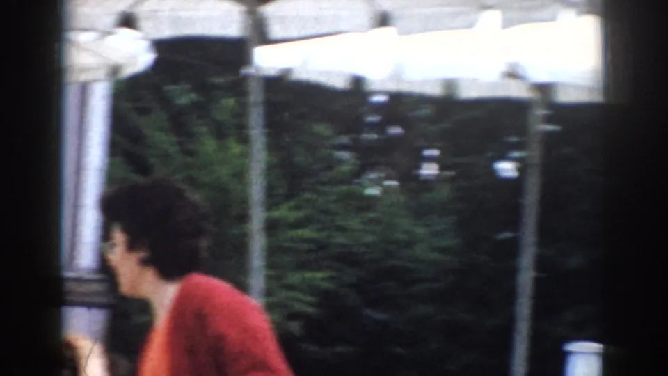 A woman in a red cardigan walks past the camera near outdoor tables and striped awnings in this 1968 Moscow home-movie frame.