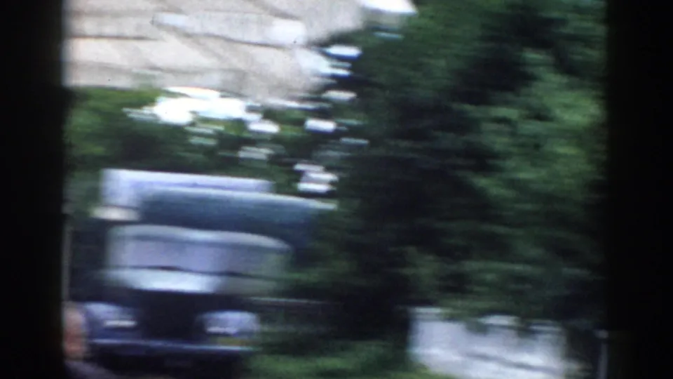 A handheld 8mm frame shows a blurred dark car beside greenery and a white balustrade in the Moscow outdoor setting.