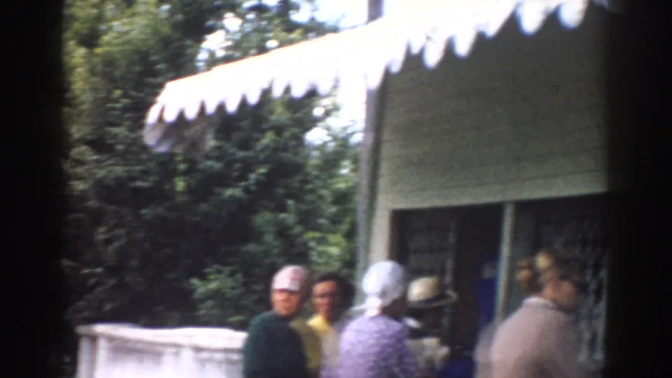 1968 Moscow home-movie frame showing adults in period clothing waiting near a restaurant or service window beneath a scalloped awning.