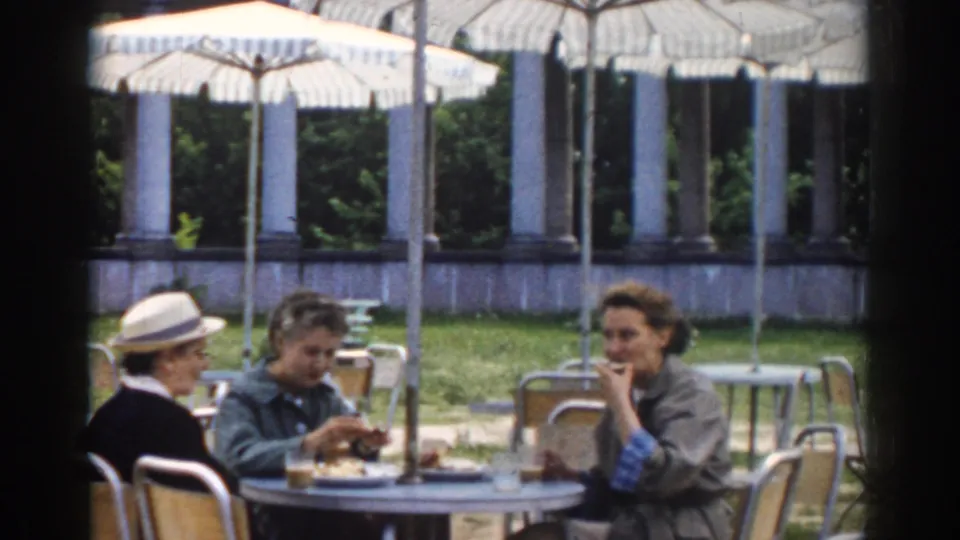 Three women at a Moscow outdoor cafe table in 1968, eating and conversing beneath striped umbrellas in a garden setting.