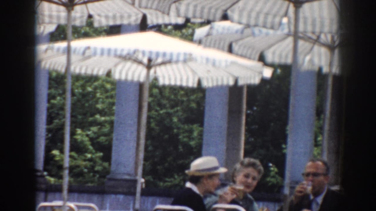 1968 Moscow home-movie frame showing adults at an outdoor café table under striped umbrellas, partly obscured by a dark vertical edge.