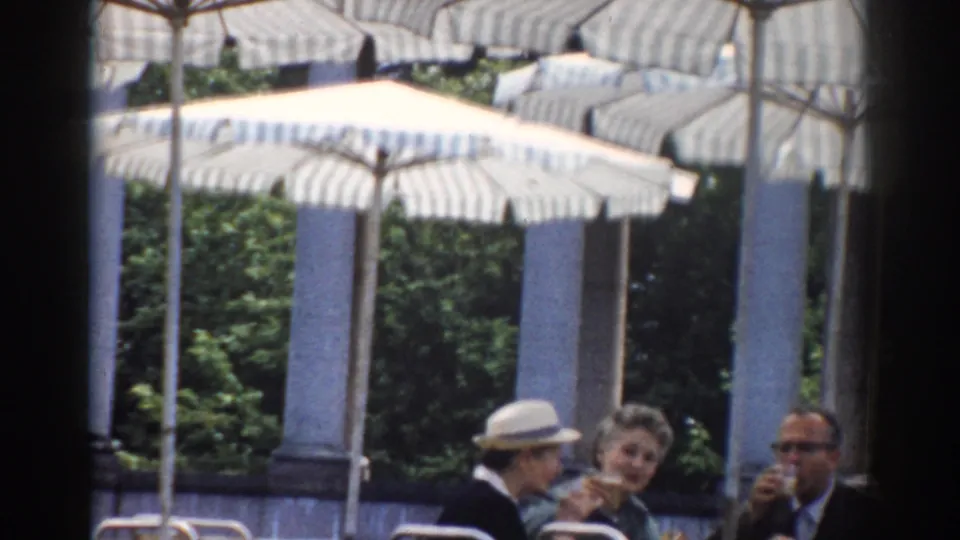 1968 Moscow home-movie frame showing adults at an outdoor café table under striped umbrellas, partly obscured by a dark vertical edge.