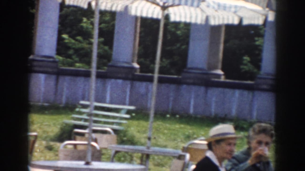 Two adults sit near café chairs and striped umbrellas with columns and greenery in the background.