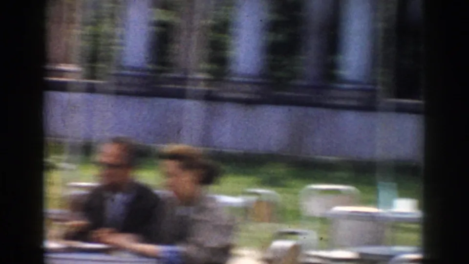 Blurred 1968 Moscow home-movie frame showing adults seated at an outdoor café, with chairs and stone columns in the background.