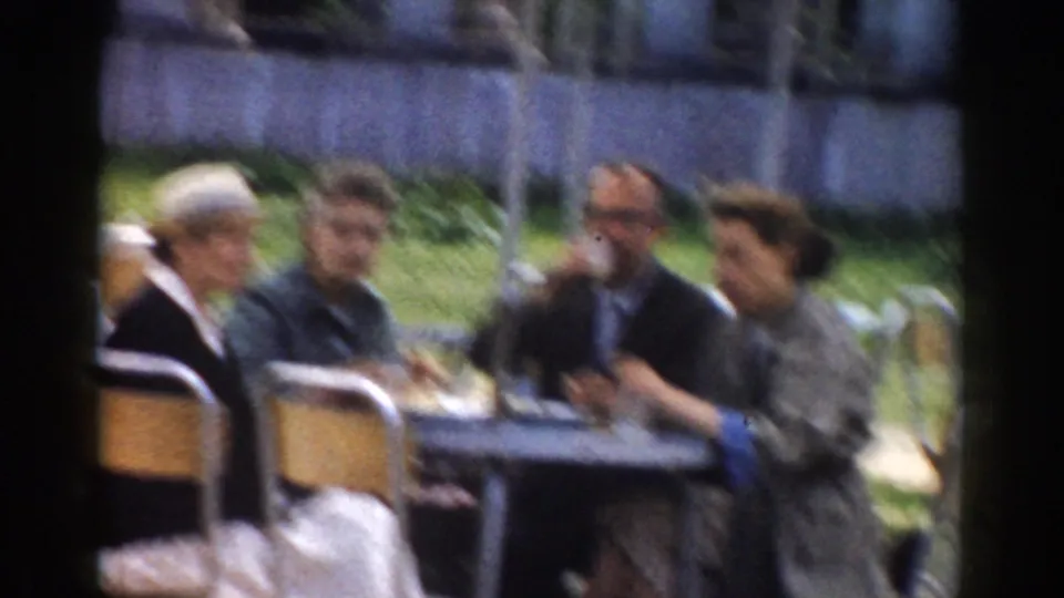 1968 Moscow home-movie frame of four adults around an outdoor café table, one in a light hat, under striped umbrellas.
