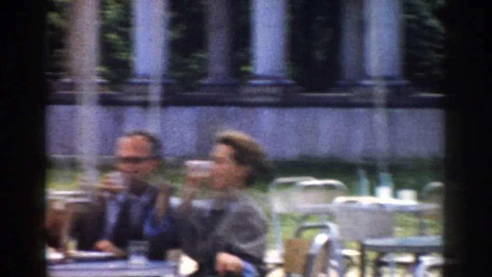 Two adults in 1968 Moscow drink at outdoor tables, with empty chairs, tree trunks, and columns visible behind them.