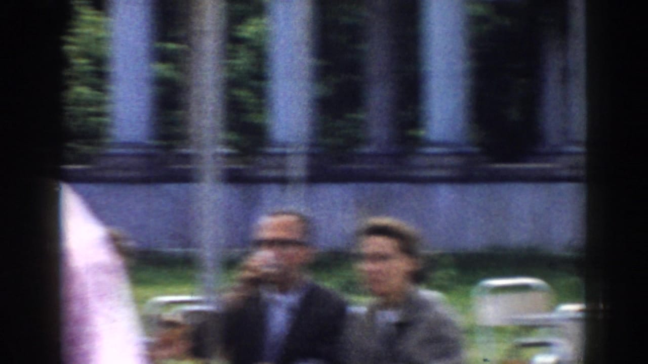 Two adults seated outdoors, one drinking, with a columned structure behind them.