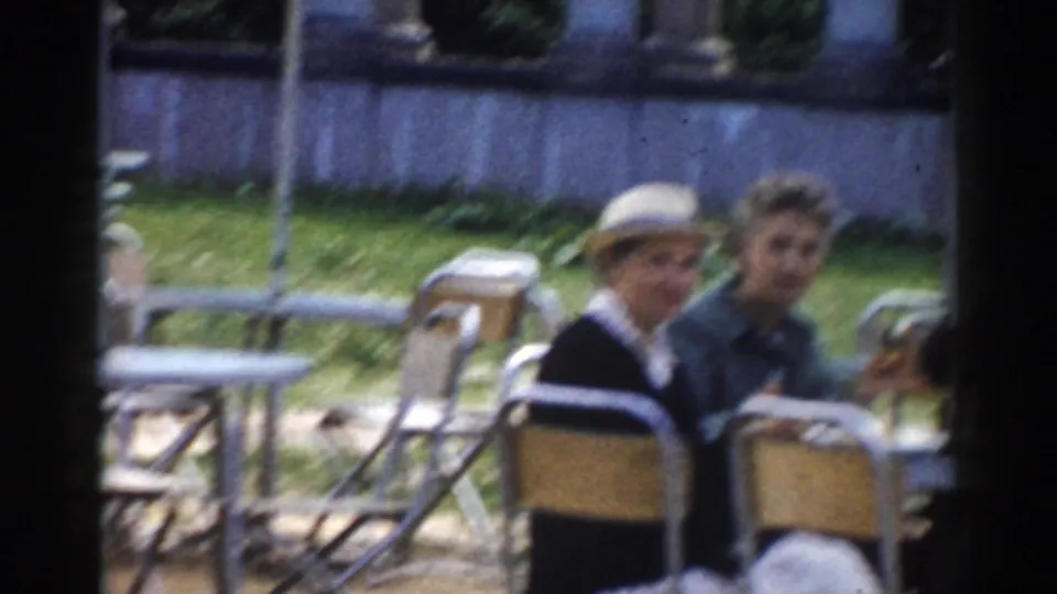 1968 Moscow home-movie frame showing two adults seated at picnic tables, with chairs, lawn, and a columned backdrop nearby.
