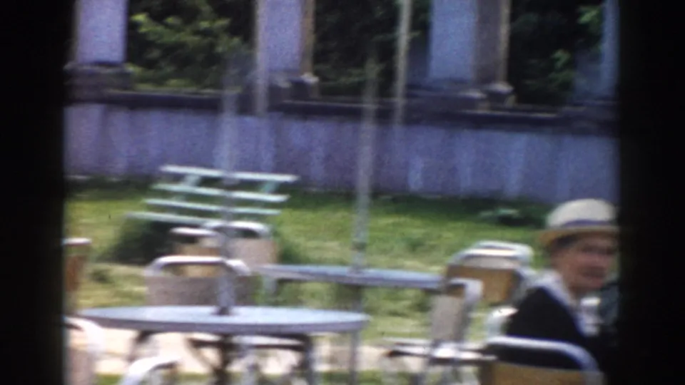 1968 Moscow home-movie frame with picnic tables, chairs, lawn, columns, and a seated adult partly visible at the right edge.