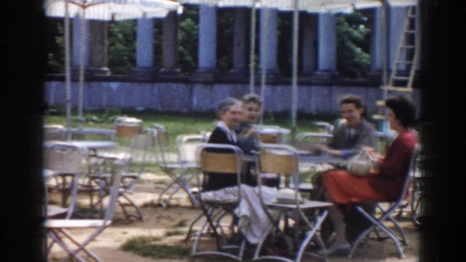 The group remains seated around the outdoor table, surrounded by empty cafe chairs and umbrellas in the Moscow courtyard setting.