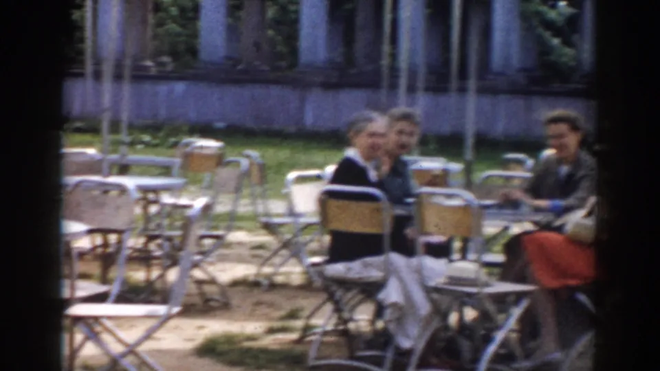 A soft 8mm frame shows four women seated at the outdoor cafe table, with chairs, umbrellas, and the columned backdrop visible.