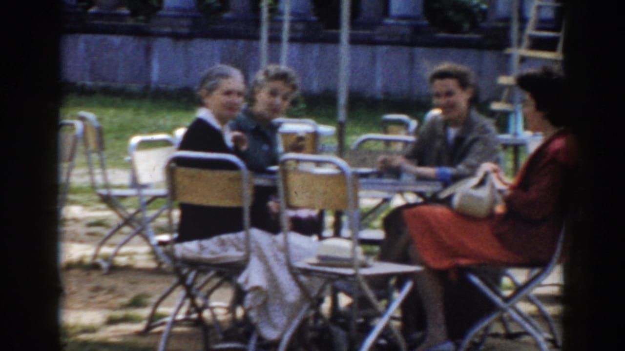 Four adults sit around outdoor cafe tables with chairs and grass behind them.