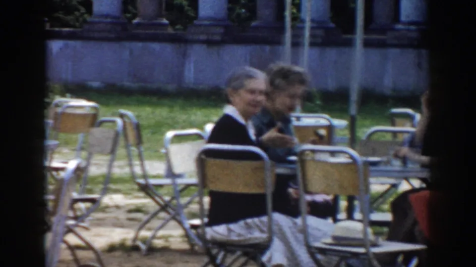 Two women at a Moscow outdoor cafe table turn close together, with empty folding chairs and a partial figure at the right edge.