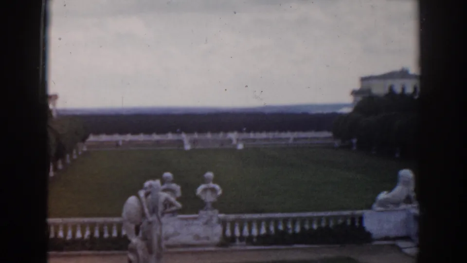 1968 Moscow home-movie frame showing terrace sculptures above a formal garden vista with lawns, hedges, trees, and palace architecture.