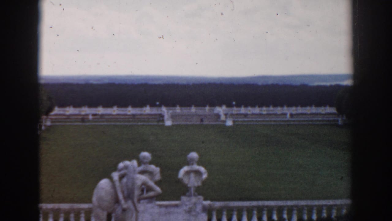 Formal garden lawn seen through dark vertical framing, with statues, white railing, distant trees, and horizon.