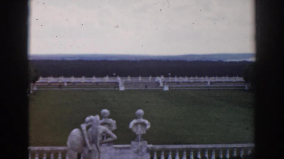 1968 Moscow home-movie view of a formal garden lawn with classical statues, a white balustrade, distant trees, and dark frame edges.