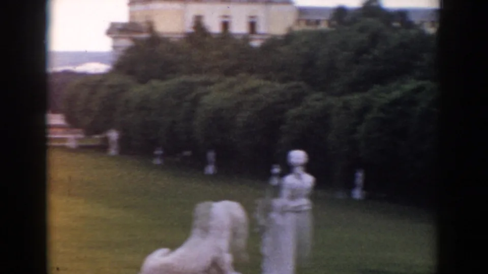 1968 Moscow home-movie frame showing clipped hedges, a classical garden statue, lawn, white fencing, and part of a large building.