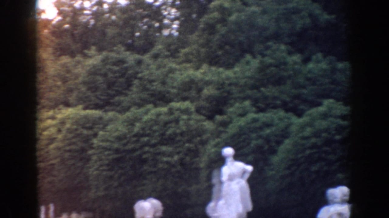 1968 Moscow home-movie frame showing white garden statues in front of dense shrubs and trees, with dark film gate edges.