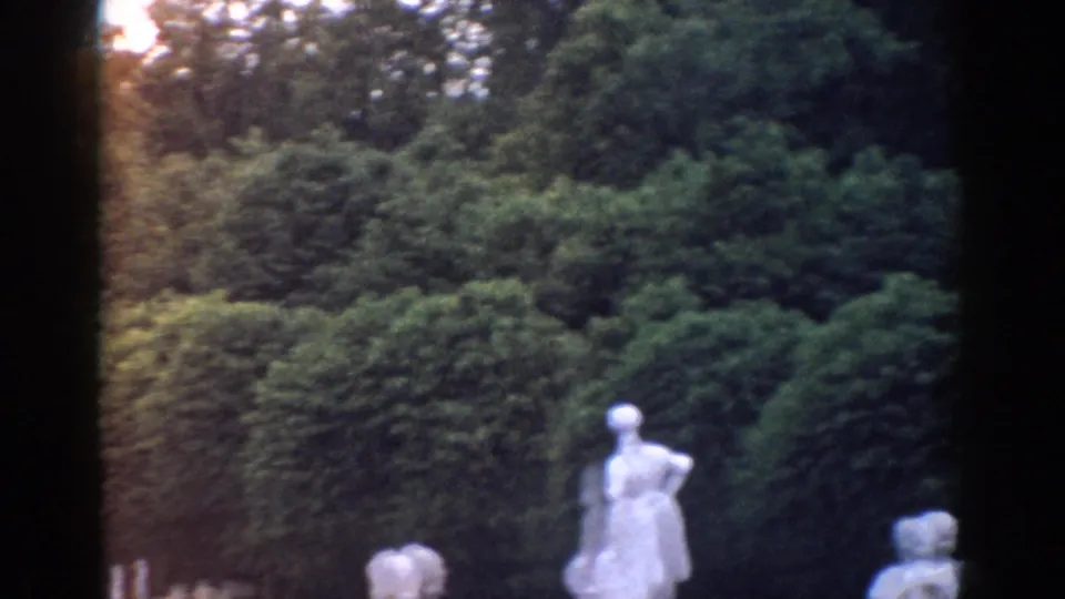 1968 Moscow home-movie frame showing white garden statues in front of dense shrubs and trees, with dark film gate edges.