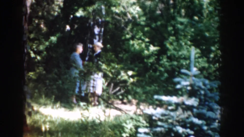 A woman in a blue dress and a man in a light hat appear to converse among trees and foliage in the 1968 Moscow home-movie footage.