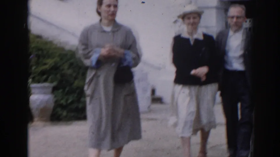 Three adults stand near a pale building wall in Moscow, with shrubs, a planter, and soft handheld 8mm focus visible.