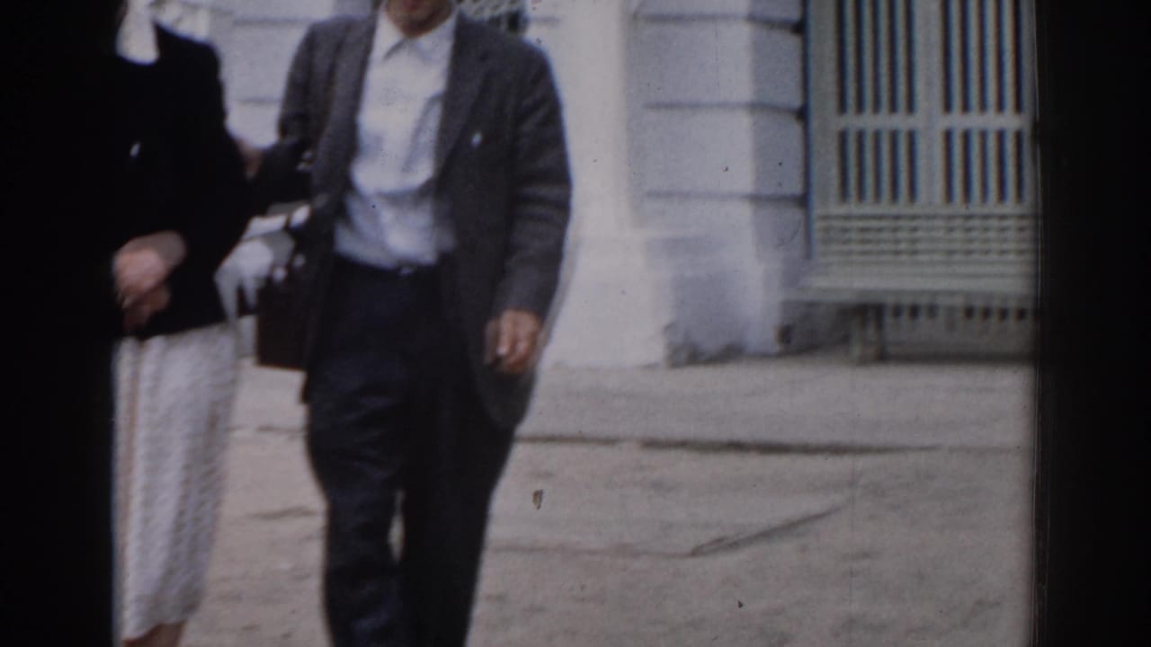 Man in glasses walking near a pale building with another figure partly visible.