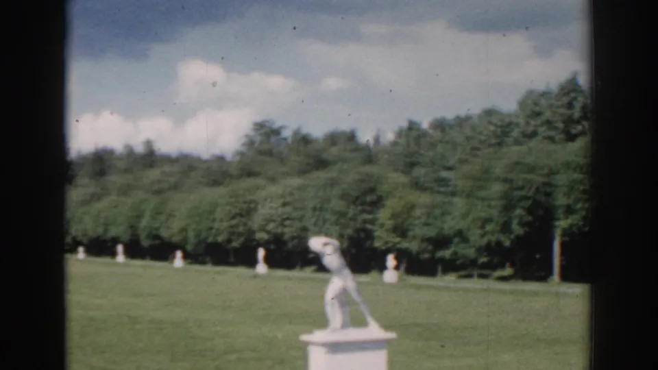A landscaped Moscow garden view shows white statues across a green lawn, backed by dense trees and a cloudy sky.