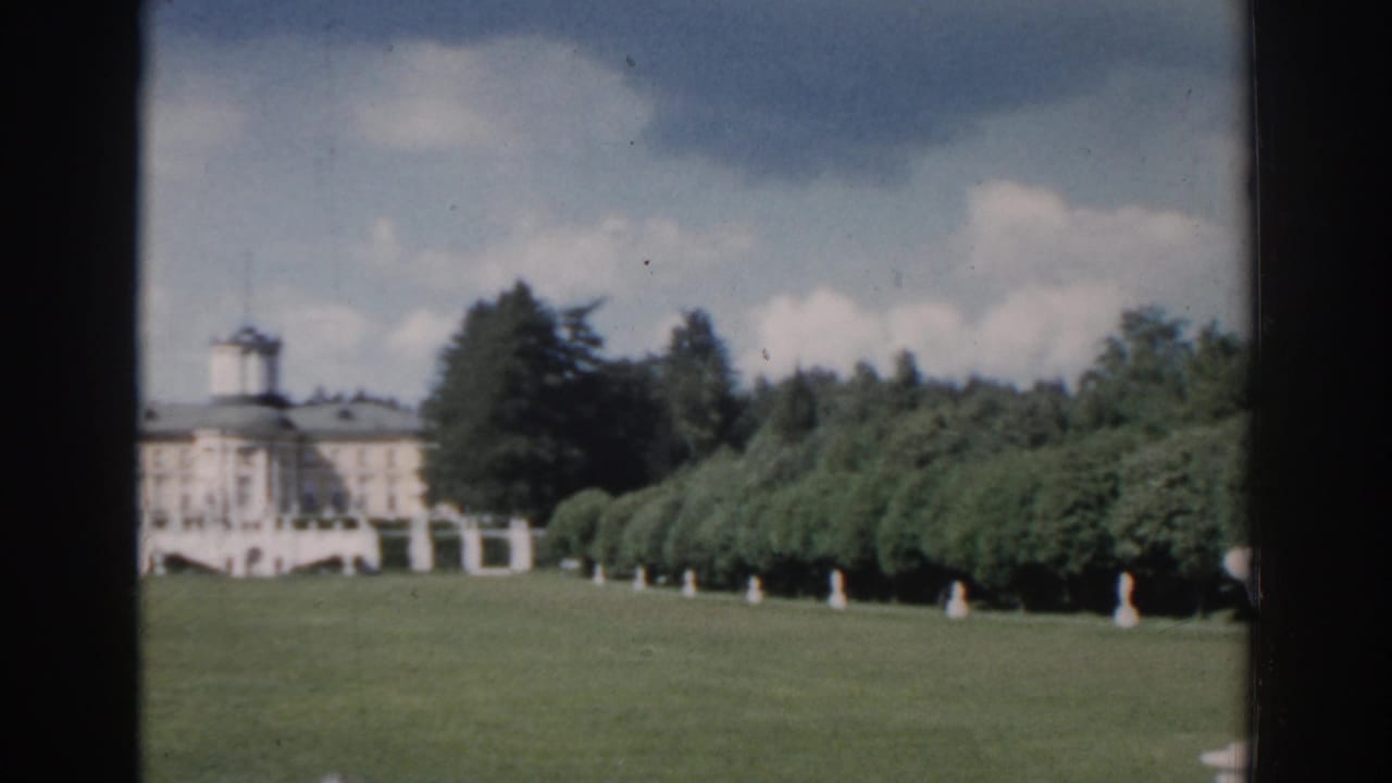 Distant mansion with central tower beyond a wide lawn and tree-lined garden.