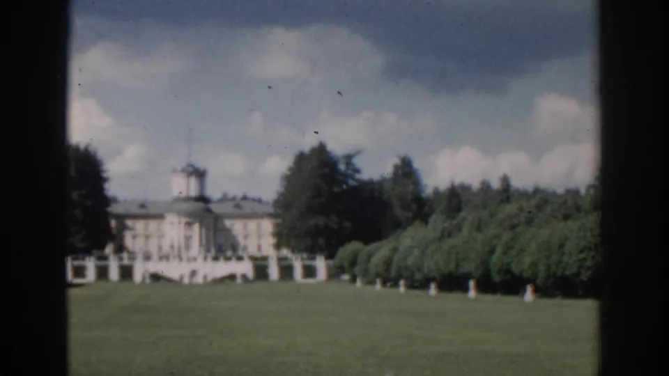 1968 Moscow 8mm frame showing the estate facade across a green lawn, with clipped trees, white statues, dark trees, and soft film grain.