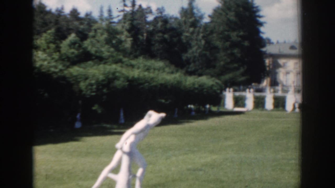 Garden lawn with a white statue, trees, and a distant building.