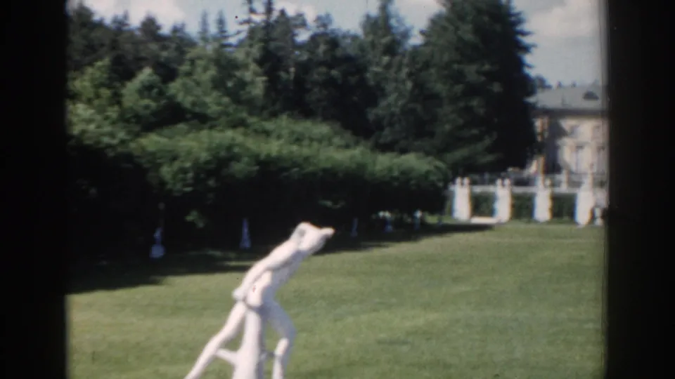 1968 Moscow home-movie frame showing a manicured garden lawn, white statue, dense trees, and a distant formal building.