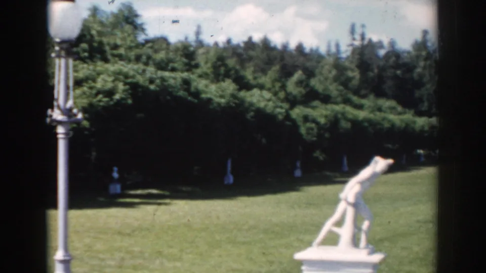 1968 Moscow home-movie frame with a lamppost in the foreground, a white garden statue, lawn, trees, and partly cloudy sky.