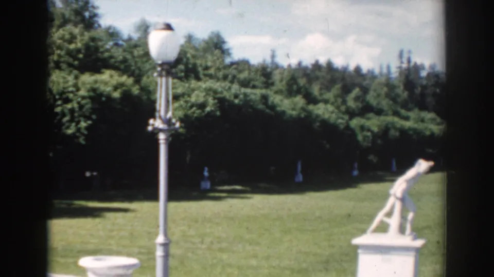 1968 Moscow home-movie frame showing a tall lamppost, a white statue on a pedestal, open lawn, and dense trees.