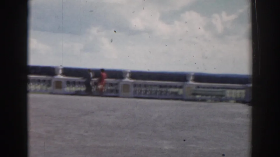 1968 Moscow home-movie frame showing a terrace or balcony railing with two distant people and a cloudy sky beyond.