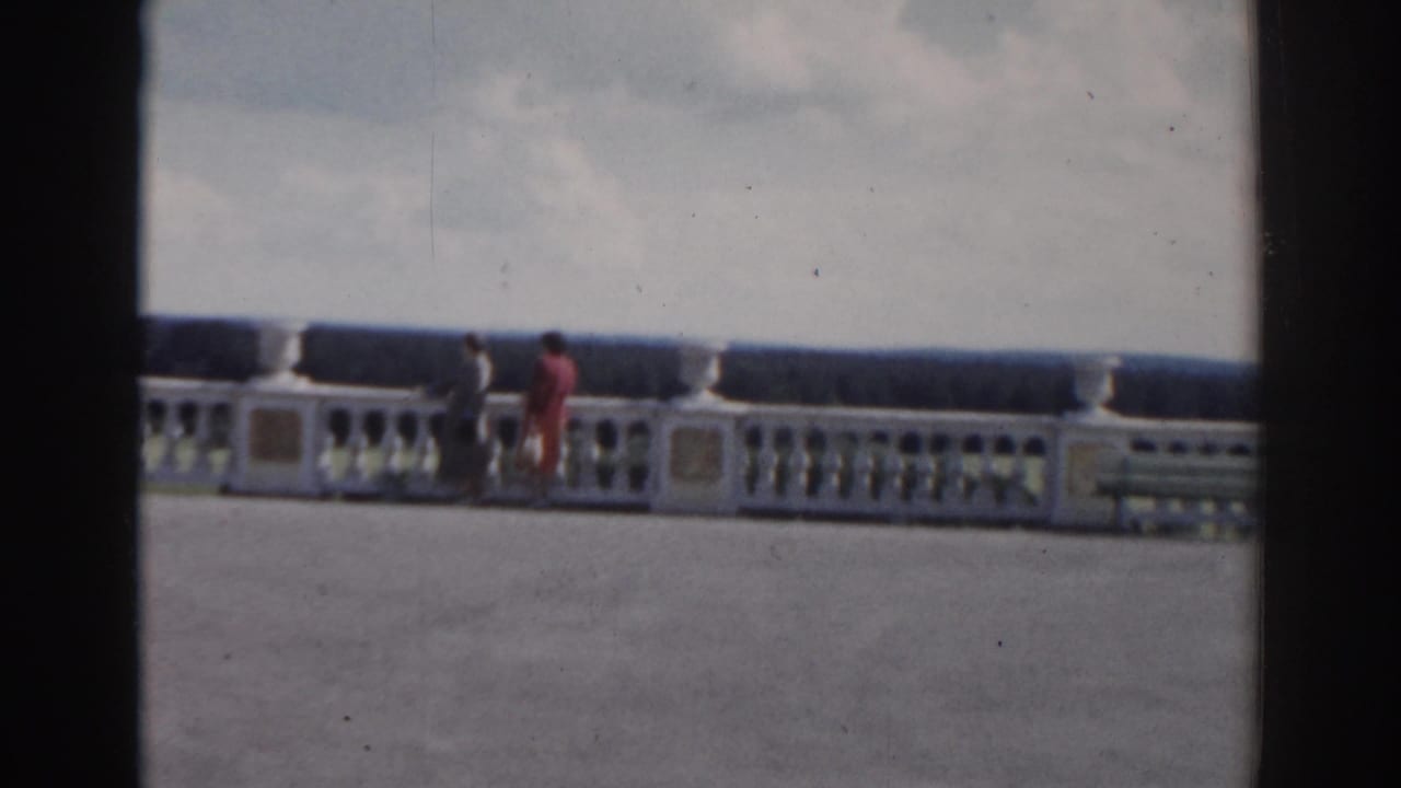Two women walk beside an ornate white balustrade on a wide terrace under a cloudy sky.