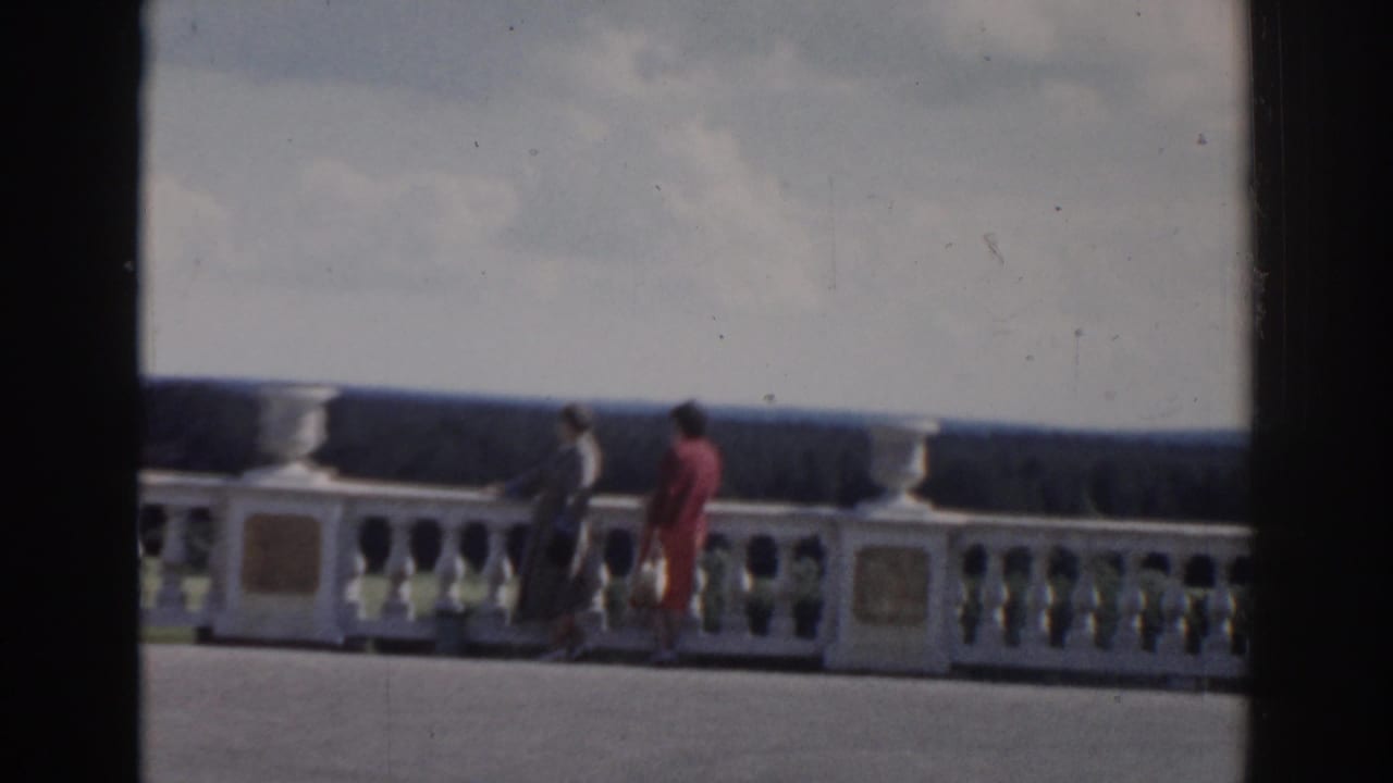Two women in 1968 Moscow footage pause or walk beside the ornate white railing, framed by cloudy sky and a distant treeline.